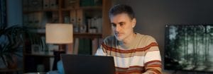 Un uomo che indossa un maglione a righe è seduto alla sua scrivania e guarda il computer portatile di fronte a lui
