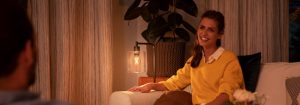 "Atmosfera accogliente nel soggiorno creata dalla luce calda della lampada LED da tavolo, che illumina una donna con maglione giallo intenta a godersi una conversazione serale."