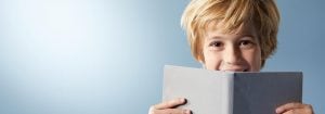"Bambino con i capelli biondi che sorride dietro un libro aperto, mostrando una gioiosa esperienza di lettura su uno sfondo blu chiaro."